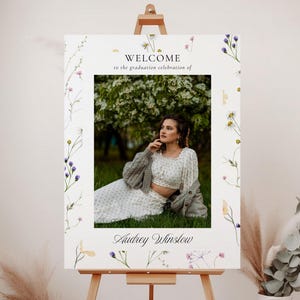 May include: Welcome to the graduation celebration of Audrey Winslow. A white sign with a floral border and a photo of a woman in a white dress and cardigan. The sign is on an easel.