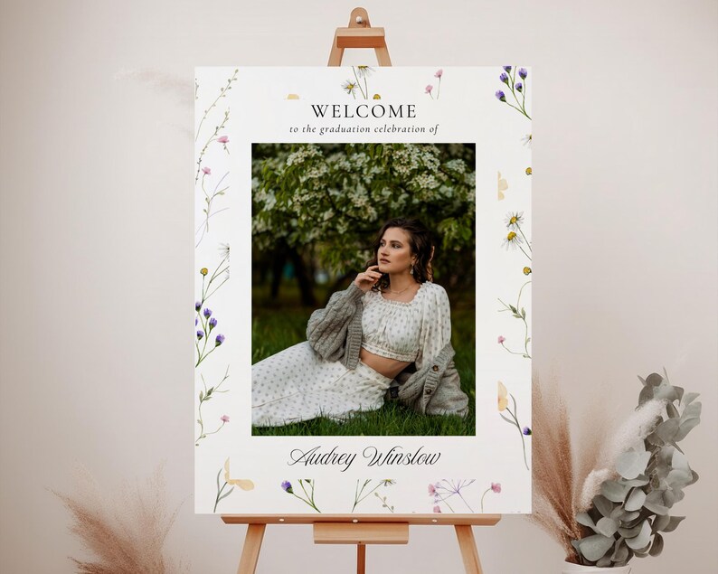 May include: Welcome to the graduation celebration of Audrey Winslow. A white sign with a floral border and a photo of a woman in a white dress and cardigan. The sign is on an easel.
