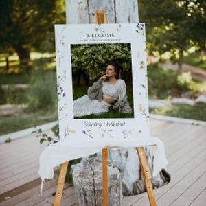 May include: A white welcome sign with a floral border and a photo of a person in a white dress and a cardigan. The sign reads "Welcome to the graduation celebration of Audrey Winslow".