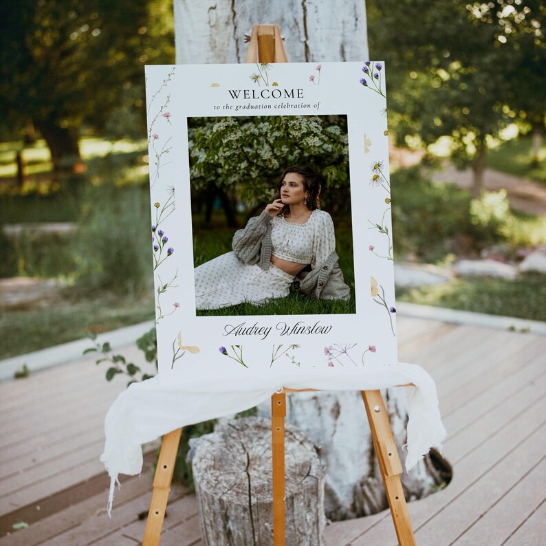 May include: A white welcome sign with a floral border and a photo of a person in a white dress and a cardigan. The sign reads "Welcome to the graduation celebration of Audrey Winslow".
