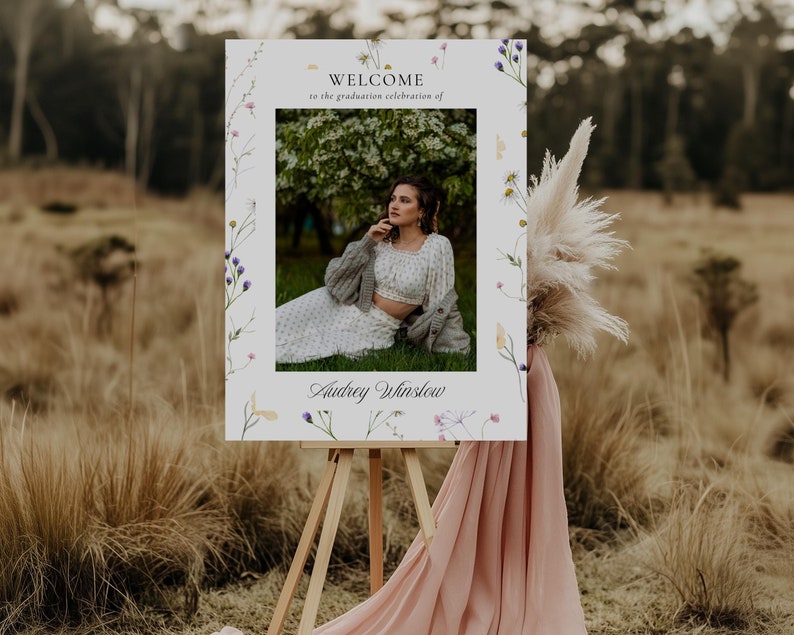 May include: A white welcome sign with a floral border and a photo of a woman in a white dress and a gray cardigan. The sign says "Welcome to the graduation celebration of Audrey Winslow".