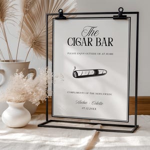 May include: A black metal frame holds a white sign that reads "The CIGAR BAR" with the text "PLEASE ENJOY OUTSIDE OR AT HOME." A drawing of a cigar is below the text. The sign is for a wedding, with the names "Archie - Colette" and a date.