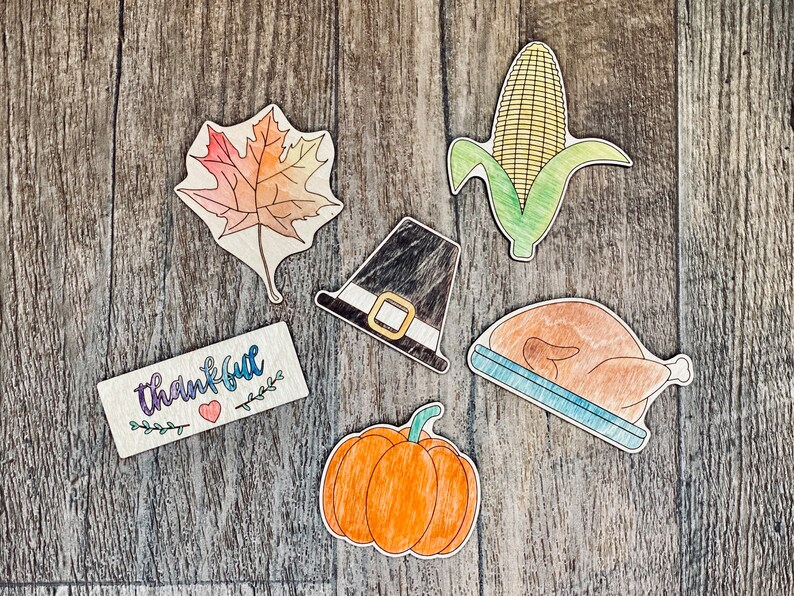 May include: A set of six wooden Thanksgiving-themed decorations. The decorations include a pilgrim hat, a cornucopia, a turkey, a pumpkin, a leaf, and a sign that says "thankful".
