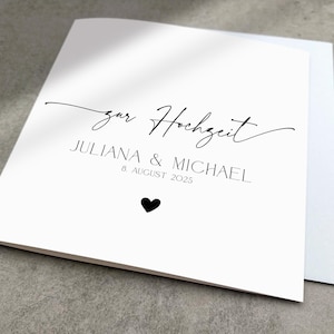 May include: A white card with black text that reads "zur Hochzeit Juliana & Michael 8 August 2025" with a small black heart below the text.