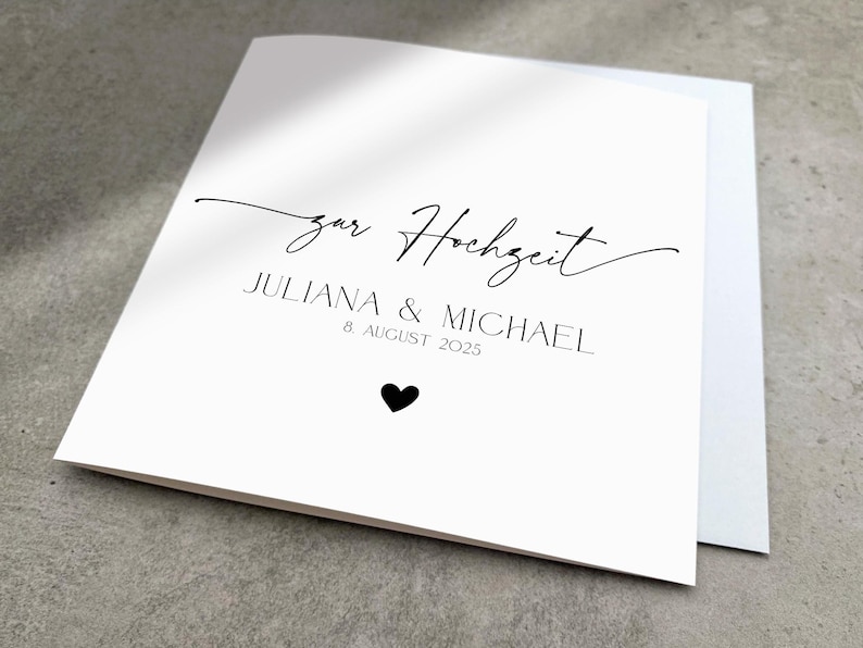 May include: A white card with black text that reads "zur Hochzeit Juliana & Michael 8 August 2025" with a small black heart below the text.