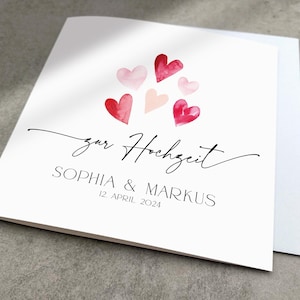 May include: A white card with a watercolor illustration of pink and red hearts. The text "zur Hochzeit" is written in black cursive script. Below the hearts, the text "Sophia & Markus" is written in black, followed by the date "12. April 2024".