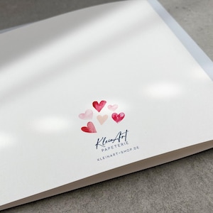 May include: A white card with a cluster of pink and red watercolor hearts. The card has the text "KleinArt Papeterie" and "kleinart-shop.de" printed in blue.