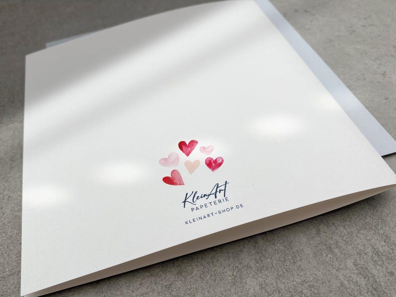 May include: A white card with a cluster of pink and red watercolor hearts. The card has the text "KleinArt Papeterie" and "kleinart-shop.de" printed in blue.