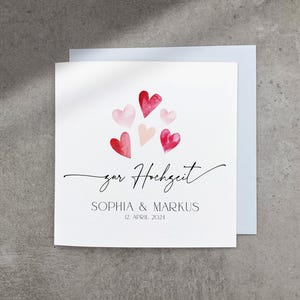 May include: A white card with a pink and red watercolor heart design. The text "zur Hochzeit" is written in cursive. Below the hearts, the text reads "Sophia & Markus 12 April 2024".