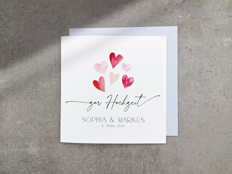 May include: A white card with a pink and red watercolor heart design. The text "zur Hochzeit" is written in cursive. Below the hearts, the text reads "Sophia & Markus 12 April 2024".