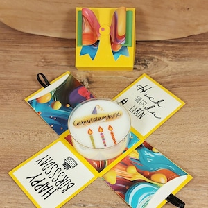 Könnte beinhalten: Eine gelbe Geschenkbox mit einer bunten, marmorierten Schleife, die sich aufklappen lässt und eine weiße Kerze in der Mitte enthüllt. Die Kerze zeigt drei Kerzen und den Text "Geburtstagskind". Die Laschen tragen die Aufschriften "Happy Bday" und "Hoch sollst du leben".