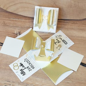 Könnte beinhalten: Eine weiße und goldene Hochzeitsgeschenkbox mit einer Schleife. Die Box ist von goldenen und weißen Karten umgeben, einige mit Text und Hochzeitsring-Grafiken. Die Box ist geöffnet und zeigt eine kleinere Box im Inneren.