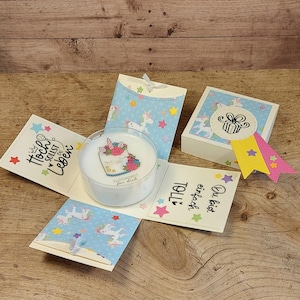 Könnte beinhalten: Eine mehrschichtige Geschenkbox mit Einhorn-Motiv. Die Box ist weiß mit blauen Akzenten und zeigt ein Einhorn-Kuchen-Design. Die Box ist von anderen Geschenkboxen und Karten mit Einhorn- und Sternmustern umgeben. Text auf Deutsch ist sichtbar.