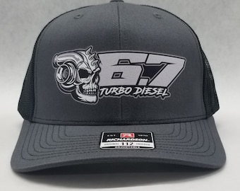 Peut inclure: Un chapeau de camionneur gris et noir avec un graphique de crâne blanc et de turbocompresseur. Le texte "6.7 TURBO DIESEL" est imprimé sur le chapeau. Le chapeau est réglable et a un dos en maille noire.