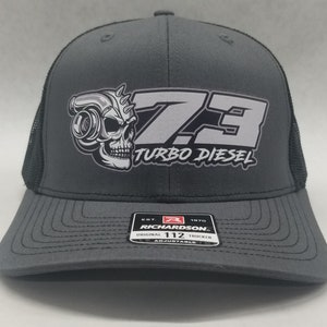 May include: Gray trucker hat with a black mesh back. The front of the hat features a white graphic of a skull with a turbocharger and the text "7.3 TURBO DIESEL". The hat is adjustable and has a Richardson 112 label.