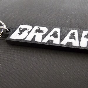 May include: Black rectangular keychain with the word "BRAAP!" in white block letters. The letters have a silhouette design inside. A silver metal split ring is attached to the keychain.