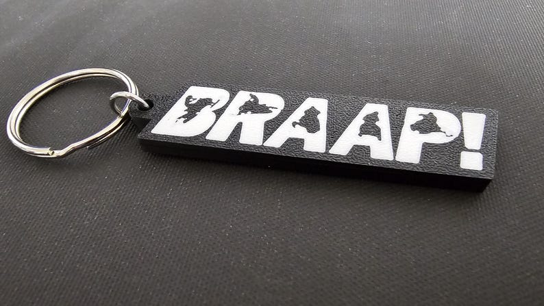 May include: Black rectangular keychain with the word "BRAAP!" in white block letters. The letters have a silhouette design inside. A silver metal split ring is attached to the keychain.