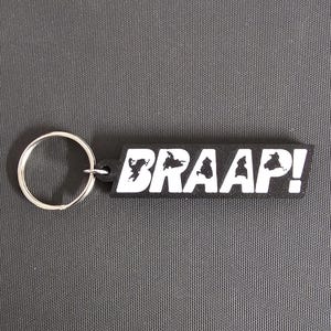 May include: Black rectangular keychain with the word "BRAAAP!" in white block letters. The keychain has a silver metal ring attached to the left side. The background is a textured gray surface.