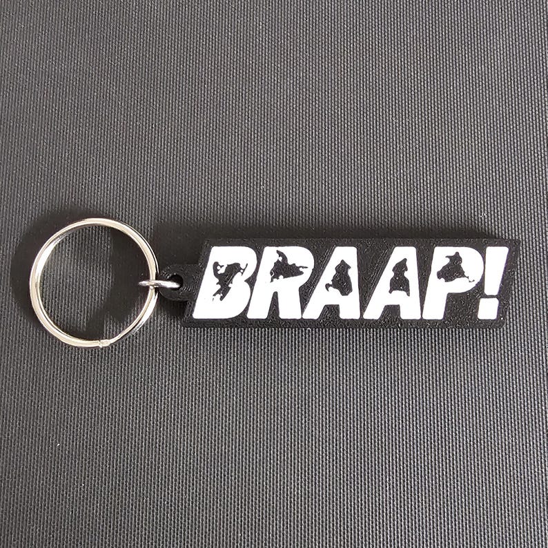 May include: Black rectangular keychain with the word "BRAAAP!" in white block letters. The keychain has a silver metal ring attached to the left side. The background is a textured gray surface.
