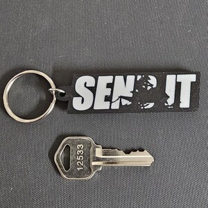 May include: A black and white keychain with the text "SEND IT" and a silver key with the number 12533. The keychain has a silver ring attached. The items are on a gray surface.
