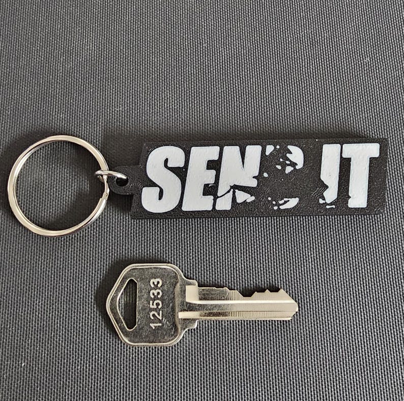 May include: A black and white keychain with the text "SEND IT" and a silver key with the number 12533. The keychain has a silver ring attached. The items are on a gray surface.