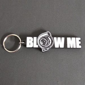 May include: Black keychain with the words "BLOW ME" in white block letters. A black and white graphic of a turbocharger is between the words. A silver key ring is attached to the left side of the keychain.