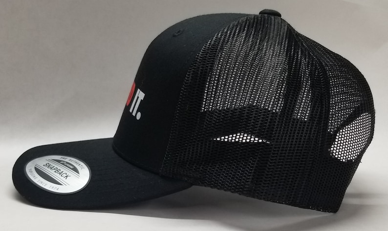 K&ouml;nnte beinhalten: Schwarze Trucker-Kappe mit einem roten Herzen und dem Text "DO IT." in Wei&szlig;. Die Kappe hat einen schwarzen Netzr&uuml;cken und ein schwarzes, gebogenes Visier.