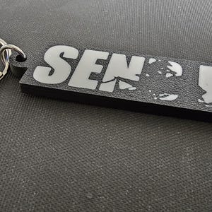 May include: Black rectangular keychain with the words "SEND IT" in white block letters. The keychain has a silver metal ring attached for keys. The background is a textured, dark gray surface.