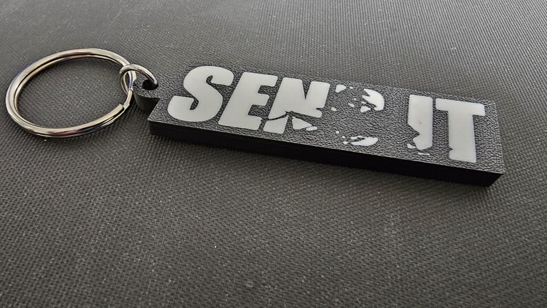 May include: Black rectangular keychain with the words "SEND IT" in white block letters. The keychain has a silver metal ring attached for keys. The background is a textured, dark gray surface.