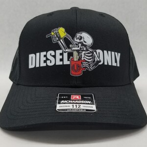 May include: Black trucker hat with a white mesh back. The front of the hat features a white graphic of a skeleton drinking from a red mug with the text "DIESEL ONLY". The hat has a black adjustable strap with a red label that reads "Richardson 112 Original Trucker Adjustable".