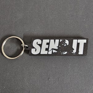 May include: Black rectangular keychain with the white text "SEND IT". The keychain has a silver key ring attached to the left side. The background is a gray textured surface.
