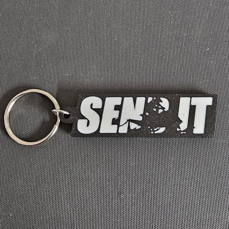 May include: Black rectangular keychain with the white text "SEND IT". The keychain has a silver key ring attached to the left side. The background is a gray textured surface.