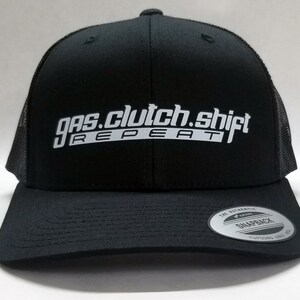 May include: Black trucker hat with a white embroidered logo that reads "gas.clutch.shift REPEAT". The hat has a black mesh back and a black snap closure.