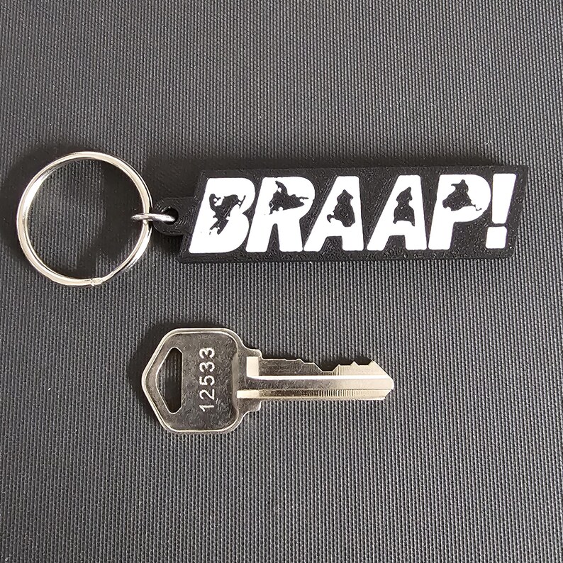 May include: A black keychain with the word "BRAAAP!" in white lettering and a silver key with the number "12533" printed on it. The keychain has a silver key ring attached.