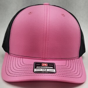 May include: A pink and black trucker hat with a black mesh back. The hat has a black stitched brim and a black label with the text "EST. 1970 RICHARDSON. ORIGINAL 112 TRUCKER ONE SIZE".