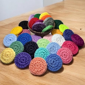May include: A collection of colorful, crocheted circular items in various shades, including yellow, blue, green, and pink. Some are arranged in a small woven basket, while others are scattered on a wooden surface. The items appear to be handmade.