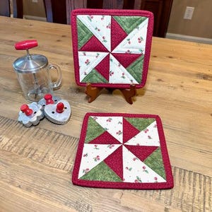 May include: Two red and green quilted pot holders with a cherry print. The pot holders are square and have a pinwheel design.