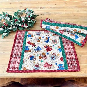 May include: A quilted Christmas placemat and runner set. The placemat features a white background with angel and star patterns, framed by a red and green border. The runner has a similar design. Festive holiday decor.