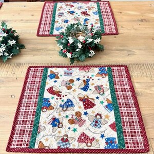 May include: Two quilted placemats with a Christmas angel theme. The placemats have a cream-colored center with angel designs in red, blue, and brown. A red and white plaid border surrounds the center, with a green stripe separating the two patterns.
