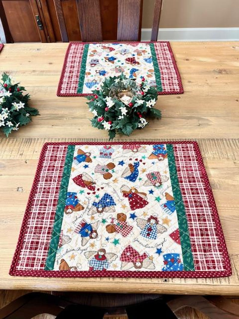 May include: Two quilted placemats with a Christmas angel theme. The placemats have a cream-colored center with angel designs in red, blue, and brown. A red and white plaid border surrounds the center, with a green stripe separating the two patterns.
