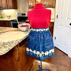 May include: A navy blue apron with a whimsical pattern of cherries and cartoon characters. The apron has two pockets and a red trim. It is displayed on a mannequin wearing a red turtleneck. The mannequin is on a blue metal stand.