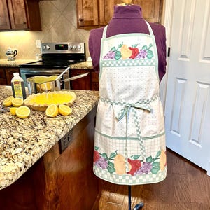 May include: A white apron with a fruit design, including pears, apples, and grapes, is displayed on a mannequin. The apron has a polka dot pattern and a green checkered border. A matching green bow ties at the waist. The kitchen setting includes a pie and lemons.