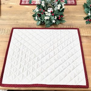 May include: A white quilted placemat with a red border featuring a small heart pattern. The placemat is on a wooden table, with a decorative wreath and a patterned table runner in the background.
