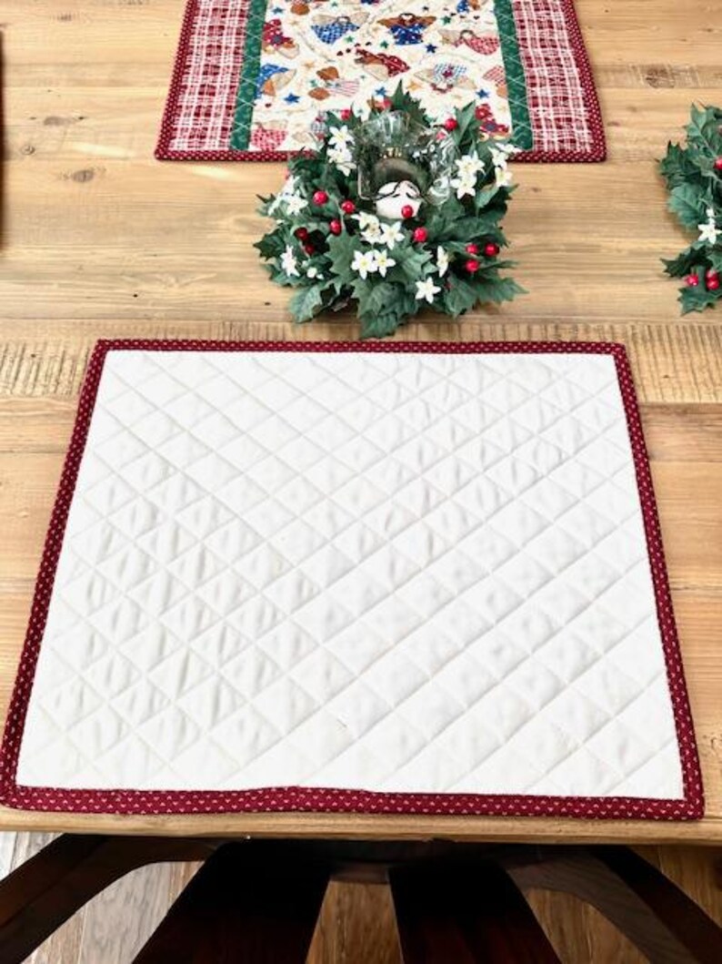 May include: A white quilted placemat with a red border featuring a small heart pattern. The placemat is on a wooden table, with a decorative wreath and a patterned table runner in the background.