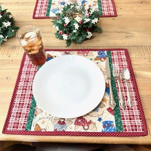 May include: A dining table setting with two placemats, each with a red and white plaid border and a central panel featuring angel figures. A white plate, silverware, and a glass of iced tea are on one placemat.