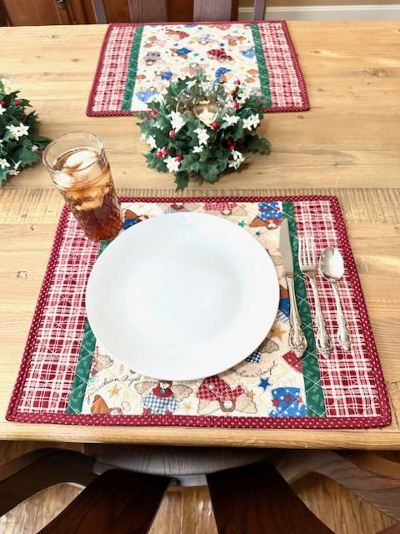 May include: A dining table setting with two placemats, each with a red and white plaid border and a central panel featuring angel figures. A white plate, silverware, and a glass of iced tea are on one placemat.