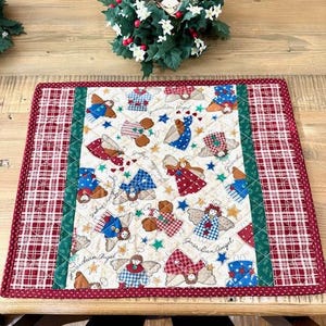 May include: Christmas-themed placemat with a quilted design. The center features a pattern of guardian angels in various outfits, stars, and the words "Guardian Angel." The border is a red and white plaid pattern.