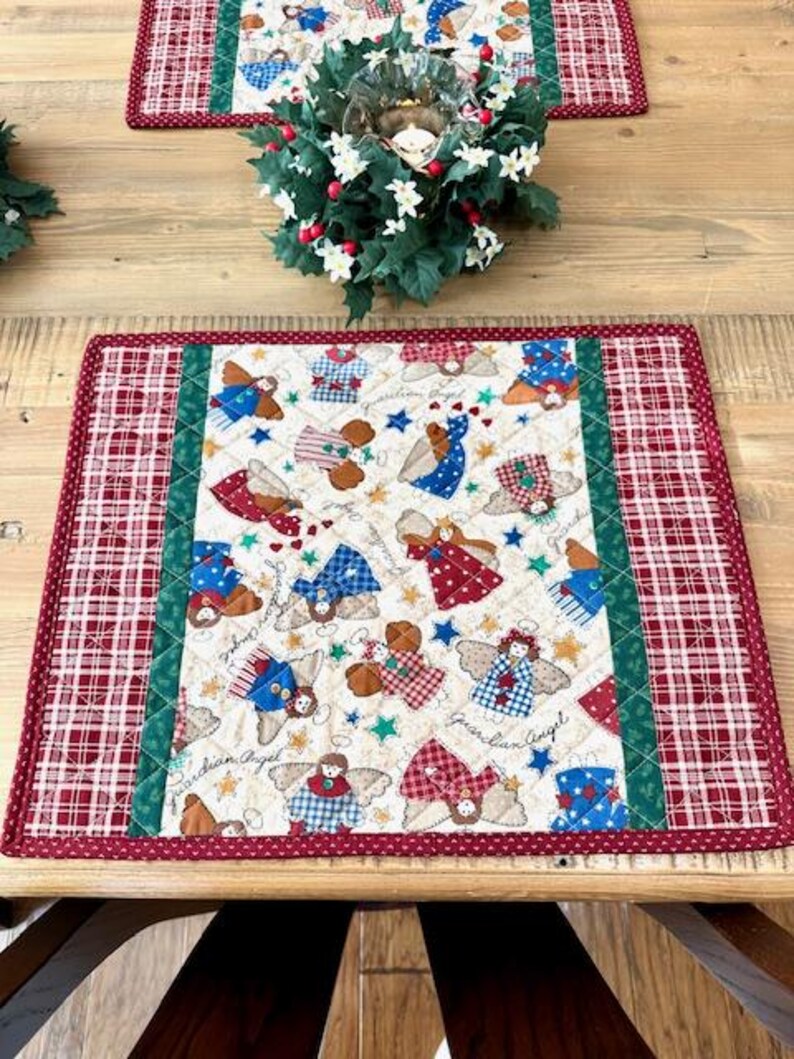 May include: Christmas-themed placemat with a quilted design. The center features a pattern of guardian angels in various outfits, stars, and the words "Guardian Angel." The border is a red and white plaid pattern.