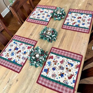 May include: A wooden table is set with four placemats and three small wreaths. The placemats have a cream-colored background with a pattern of angels, stars, and gifts, framed by a red plaid border. The wreaths are green with white berries.