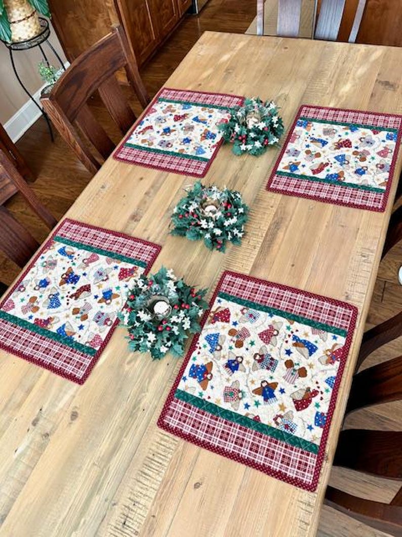 May include: A wooden table is set with four placemats and three small wreaths. The placemats have a cream-colored background with a pattern of angels, stars, and gifts, framed by a red plaid border. The wreaths are green with white berries.